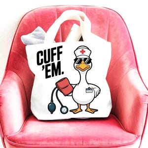 May include: White tote bag with black text that reads "CUFF 'EM." The bag features a cartoon goose wearing a nurse's hat and sunglasses, holding a blood pressure cuff. The bag has white handles and is sitting on a pink chair.