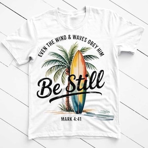May include: White t-shirt featuring a colorful surfboard design with a palm tree and the words "Be Still." The text "Even the wind & waves obey him" is arched above the surfboard, with "Mark 4:41" below. The shirt has a crew neck.
