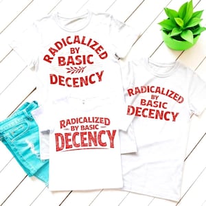 Op de afbeelding: Witte t-shirts met de tekst "RADICALIZED BY BASIC DECENCY" in het rood. De shirts zijn gerangschikt op een wit houten oppervlak met een spijkerbroek en een kleine groene plant in een pot.