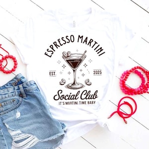 May include: White t-shirt with an "Espresso Martini Social Club" graphic. The design features a martini glass, coffee beans, and the text "It's Martini Time Baby". The shirt is paired with denim shorts and red beaded bracelets.