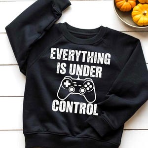 Puede incluir: Sudadera negra con texto blanco que dice "EVERYTHING IS UNDER CONTROL" con un gráfico de un mando de videojuegos. La sudadera es de cuello redondo.