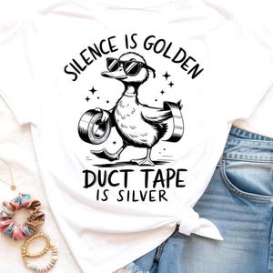 May include: White t-shirt featuring a cartoon duck wearing sunglasses and carrying duct tape. The text "SILENCE IS GOLDEN DUCT TAPE IS SILVER" is printed above and below the duck illustration in black.