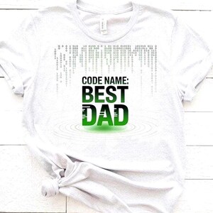 May include: A light gray t-shirt with the text "CODE NAME: BEST DAD" in black and green. The design includes a binary code pattern and water ripple effect. A perfect gift for Father's Day.