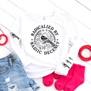 Op de afbeelding: Wit shirt met lange mouwen met een cirkelvormige afbeelding met de tekst "RADICALIED BY BASIC DECENCY" rond een vogelillustratie. Extra vogelafbeeldingen staan op de mouwen. De outfit wordt gecompleteerd door een spijkerbroekshort en roze sokken.