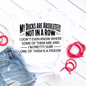 Puede incluir: Camiseta blanca con el texto "MY DUCKS ARE ABSOLUTELY NOT IN A ROW". La camiseta se combina con pantalones cortos de mezclilla y pulseras de cuentas rojas. El texto dice con humor: "Ni siquiera sé dónde están algunos de ellos y, estoy bastante seguro, uno de ellos es una paloma."