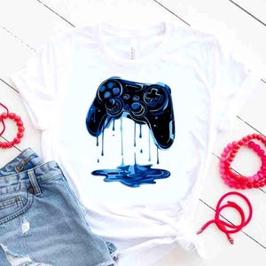 May include: White t-shirt featuring a blue and black video game controller design. The controller has a dripping effect, with blue liquid forming a puddle at the bottom. The shirt is paired with denim shorts and red beaded jewelry.