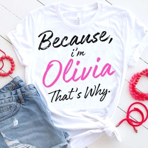 May include: White t-shirt with the text "Because, I'm Olivia That's Why." in black and pink lettering. The shirt is paired with denim shorts and red bracelets.