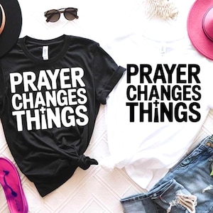 Peut inclure: Deux t-shirts, un noir et un blanc, avec l'inscription "PRAYER CHANGES THINGS" en lettres blanches en gras. Le t-shirt noir est noué. Des lunettes de soleil, un chapeau rose et un short en jean sont également visibles.