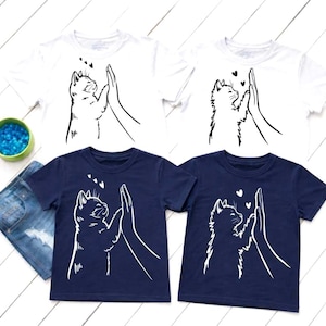 May include: Four short-sleeved t-shirts, two white and two navy blue, are displayed. Each shirt features a black and white graphic of a cat high-fiving a hand, with small heart details. The shirts are likely for children.