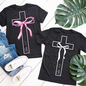 May include: Two black t-shirts with a white cross and a pink ribbon on one shirt and a white ribbon on the other. The text "John 3:16" is written on both shirts.
