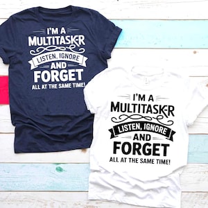 May include: Two t-shirts, one navy blue and one white, with the same humorous text in black. The text reads "I'M A MULTITASKER, LISTEN, IGNORE AND FORGET ALL AT THE SAME TIME!" The shirts are on a light blue and wood-toned surface.