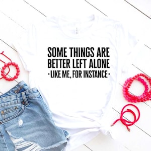 May include: White t-shirt with the text "SOME THINGS ARE BETTER LEFT ALONE - LIKE ME, FOR INSTANCE-" in black. The shirt is paired with denim shorts and red beaded bracelets. The items are arranged on a white wooden surface.