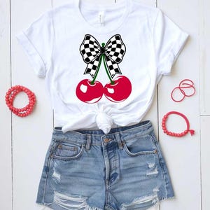 May include: White t-shirt with a black and white checkered bow on top of two red cherries. The shirt is worn with light blue denim shorts.