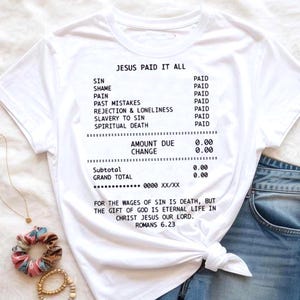 May include: White t-shirt with the text "JESUS PAID IT ALL" printed in black, resembling a receipt. The receipt lists various sins and states they are "PAID". The bottom of the shirt includes a Bible verse from Romans 6:23.