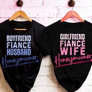 以下が含まれることがあります： 木製のハンガーに掛けられた2枚の黒いTシャツ。1枚のシャツには、白と青で「BOYFRIEND FIANCE HUSBAND Honeymooner」と書かれています。もう1枚のシャツには、白とピンクで「GIRLFRIEND FIANCE WIFE Honeymooner」と書かれています。