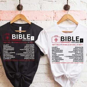 May include: Two white t-shirts with black text. The text reads "BIBLE EMERGENCY HOTLINES GET HELP + ANGELS ON CALL 24/7" and lists bible verses for different situations like hurt, sin, betrayal, stress, sadness, lost hope, anger, worry, being in danger, and loneliness. One shirt is black and the other is white.