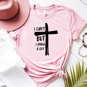 May include: A pink t-shirt with a black cross and the text "I can't but I know a guy" printed on it.