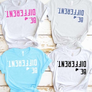 Puede incluir: Cuatro camisetas con la frase "BE. DIFFERENT." impresa. Las camisetas son blancas, azul claro y gris claro. El texto es rosa, azul marino, blanco y negro. Cada camiseta tiene un pequeño símbolo de corazón.