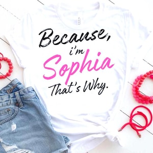 May include: White t-shirt with the text "Because, I'm Sophia That's Why" in black and pink lettering. The shirt is paired with denim shorts and red bracelets. The shirt is knotted at the waist.