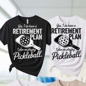 May include: Black and white t-shirts with the text "Yes, I do have a RETIREMENT PLAN. I plan on playing Pickleball." A graphic of a pickleball and paddle is also featured. The shirts are tied at the waist.