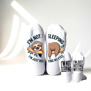 May include: White socks featuring a cartoon sloth and the text "I'M NOT SLEEPING, I'M JUST RESTING MY EYES." The sloth is brown and resting on a branch. Two smaller socks with the same text are also visible.