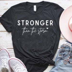 May include: Black t-shirt with white text that reads "Stronger than the storm" with a small heart symbol.