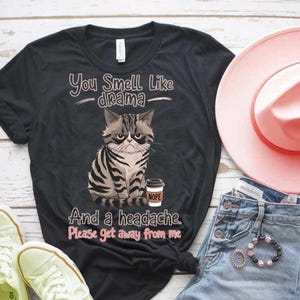 May include: Black t-shirt with a grumpy cat illustration holding a coffee cup that says "NOPE". The text on the shirt reads "You Smell Like -drama- And a headache. Please get away from me."