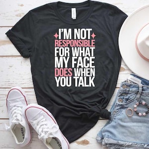 Könnte beinhalten: Schwarzes T-Shirt mit weißem Text, der "Ich bin nicht verantwortlich für das, was mein Gesicht tut, wenn du sprichst" lautet. Der Text ist in Pink umrandet. Das Shirt liegt auf einer weißen Holzoberfläche mit einem Paar weißen Sneakers, einer Jeans-Shorts und einem weißen Hut.