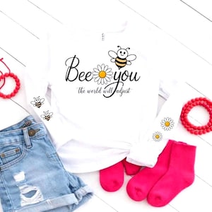 Puede incluir: Camiseta blanca de manga larga con el texto "Bee you the world will adjust" y gráficos de abejas y margaritas. La imagen también incluye pantalones cortos de mezclilla, calcetines rosas y pulseras de cuentas rojas.