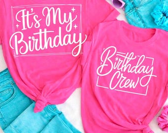 Może przedstawiać: Dwie jaskraworóżowe koszulki z białym napisem. Na jednej widnieje napis "It's My Birthday", a na drugiej "Birthday Crew". Na zdjęciu znajdują się również jasnoniebieskie dżinsy, fioletowe okulary przeciwsłoneczne i różowa gumka do włosów. Koszulki są zawiązane na dole.