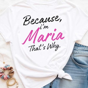 Könnte beinhalten: Weißes T-Shirt mit dem Text "Because, I'm Maria That's Why." Das Wort "Maria" ist in einer leuchtend rosa Schrift geschrieben, während der Rest des Textes schwarz ist. Das T-Shirt ist an der Taille geknotet.