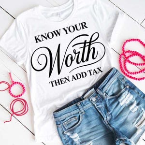 May include: A white t-shirt with the text "KNOW YOUR WORTH THEN ADD TAX" in black script. The shirt is paired with denim shorts and pink beaded bracelets. The image is taken on a white wooden surface.
