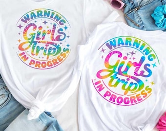 May include: Two white t-shirts with the text "WARNING Girls Trip IN PROGRESS" in a rainbow tie-dye design. The shirts are paired with denim shorts, sunglasses, and scrunchies, suggesting a vacation theme.