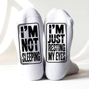 以下が含まれることがあります： 黒い文字が入った白い靴下のペア。片方の靴下には「I'M NOT SLEEPING」、もう片方には「I'M JUST RESTING MY EYES」と書かれています。柔らかく快適な素材で作られています。