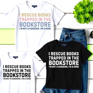 May include: Three t-shirts with the text "I RESCUE BOOKS TRAPPED IN THE BOOKSTORE. I'M NOT A HOARDER, I'M A HERO." One white, one black, and one white with colored text. The shirts are paired with blue jeans.