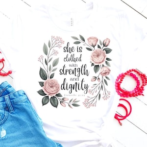 May include: White t-shirt featuring the quote "she is clothed with strength and dignity" surrounded by a floral wreath of pink roses and green leaves. The text is in a stylish script font. The shirt is paired with blue jeans and red accessories.