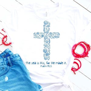 May include: White t-shirt with a blue cross design filled with seashell illustrations. The text "the sea is His, for He made it. Psalm 95:5" is printed below the cross. The shirt is paired with denim shorts and red bracelets.