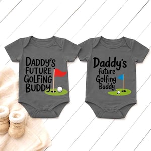 May include: Two gray baby onesies with the text "Daddy's Future Golfing Buddy" and a golf-themed graphic. One has a red flag, the other a blue flag. A golf ball and green putting green are also featured.