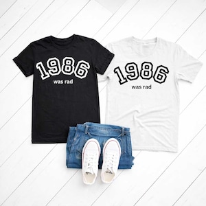 Puede incluir: Dos camisetas, una negra y otra blanca, con el texto "1986 was rad" en negrita y con contorno. Las camisetas se muestran con vaqueros azules y zapatillas blancas sobre una superficie de madera blanca.