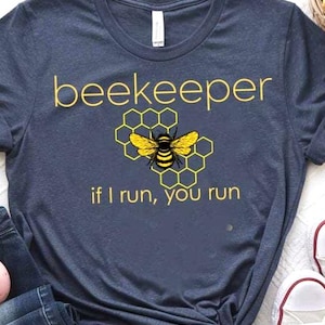 May include: Navy blue t-shirt with the word "beekeeper" in yellow, along with a yellow and black bee graphic and the phrase "if I run, you run." The shirt is styled with jeans and white sneakers.