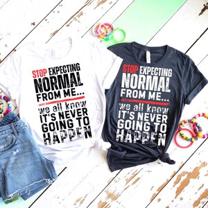 May include: Two t-shirts, one white and one dark gray, with the text "STOP EXPECTING NORMAL FROM ME... we all know IT'S NEVER GOING TO HAPPEN" in black and red. The shirts are paired with denim shorts and colorful bracelets.