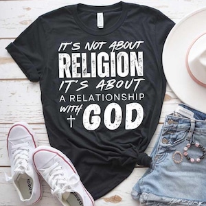 Może przedstawiać: Czarny T-shirt z białym tekstem, który brzmi "Nie chodzi o religię, chodzi o relację z Bogiem" z symbolem krzyża. Koszulka leży na drewnianej powierzchni z parą białych butów sportowych, dżinsowymi spodenkami i białym kapeluszem.