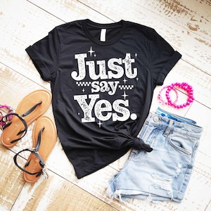 Puede incluir: Camiseta negra con el texto blanco "Just say Yes", combinada con shorts vaqueros, sandalias y una pulsera de cuentas rosas. La camiseta está anudada a la cintura, creando un look casual. Las sandalias son marrones con correas negras.