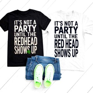 Op de afbeelding: Zwarte en witte t-shirts met de tekst "IT'S NOT A PARTY UNTIL THE REDHEAD SHOWS UP". Het zwarte shirt heeft een groene klaver. Jeans en neon groene sneakers zijn ook zichtbaar.