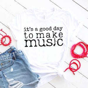 May include: White t-shirt with the text "it's a good day to make music" in black. The shirt is paired with denim shorts and red beaded bracelets. The image is taken on a white wooden surface.