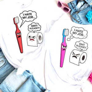Puede incluir: Dos camisetas blancas con gráficos de dibujos animados humorísticos. Una presenta un cepillo de dientes rojo y la otra un cepillo de dientes rosa, ambos con caras enfadadas y bocadillos que dicen "I HATE MY JOB..." y "OH!!! PLEASE!!!".