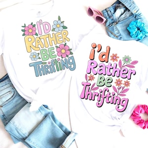 Op de afbeelding: Twee witte T-shirts met de tekst "I'd Rather Be Thrifting" in kleurrijke letters, omringd door bloemmotieven. De shirts worden gecombineerd met een blauwe jeans, zonnebril en scrunchies.