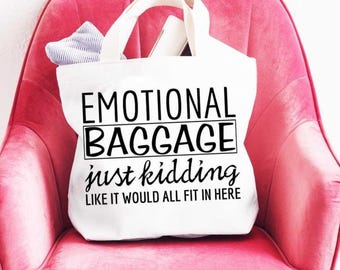 Op de afbeelding: Witte canvas draagtas met zwarte tekst die "Emotional Baggage just kidding like it would all fit in here" zegt. De tas ligt op een roze stoel.