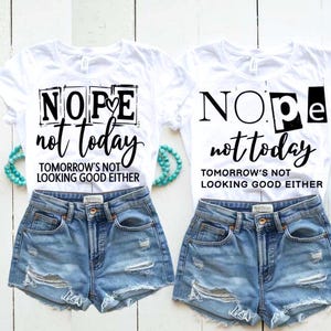 以下が含まれることがあります： 「NOPE not today TOMORROW'S NOT LOOKING GOOD EITHER」の黒い文字が入った白いTシャツ2枚。ダメージ加工のデニムショートパンツと組み合わせています。ターコイズ色のビーズブレスレットがシャツの近くに置かれています。
