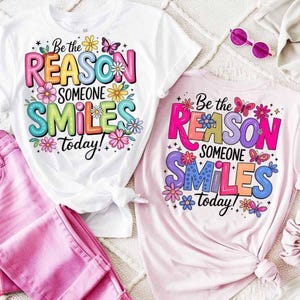 以下が含まれることがあります： 「Be the REASON SOMEONE SMILES today!」とカラフルな文字で書かれた、白とピンクの2枚のTシャツ。デザインには花と蝶が含まれています。ピンクのパンツとサングラスも写っています。